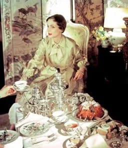 Vintage scene of a woman in elegant attire serving tea in a richly decorated room with silver tea set and cake on the table.