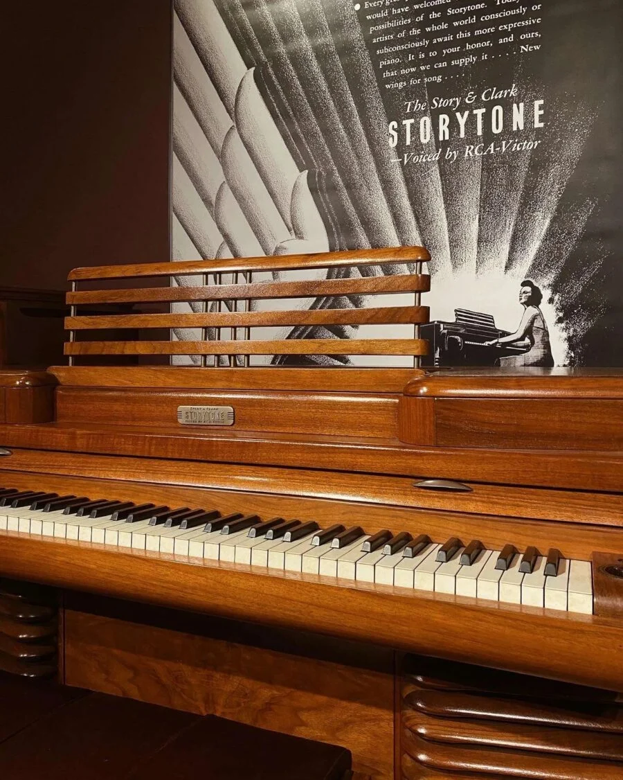 Close-up of a vintage wooden piano with Storytone branding and a mural of a woman playing a piano on the wall behind it.