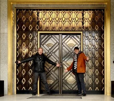 Two men standing in front of an ornate Art Deco door with gold and bronze geometric patterns