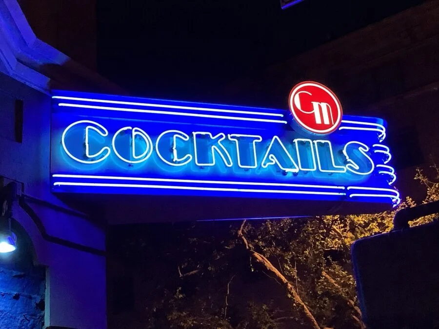 Blue neon sign glowing at night reading 'Cocktails' with a red circular logo above it.