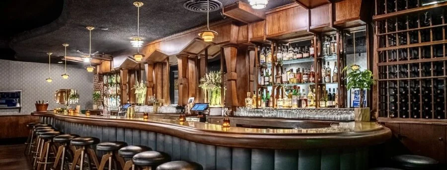 Interior view of a stylish bar with wooden decor, leather bar stools, and bottles displayed behind the counter under warm lighting.