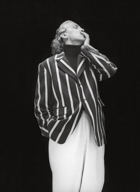 Black and white photo of a person wearing a vertically striped blazer and white pants, posing with one hand touching their face and the other in a pocket.
