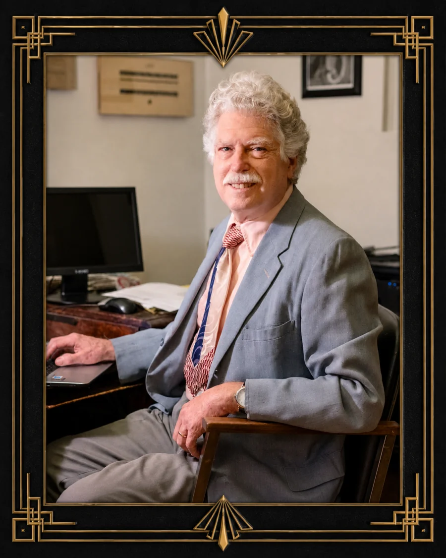 Portrait of a smiling elderly man with curly white hair and a mustache, wearing a light gray suit and a colorful tie, sitting at a desk with a computer in the background.