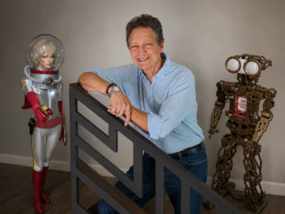 Man smiling and leaning on a black metal railing with a robot in a silver space suit on the left and a bronze mechanical robot on the right