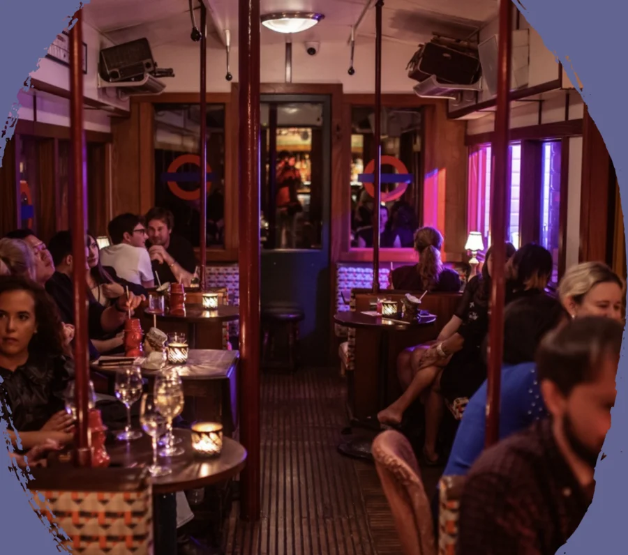 Interior of a cozy, dimly lit bar or café with people seated at small tables and warm ambient lighting.