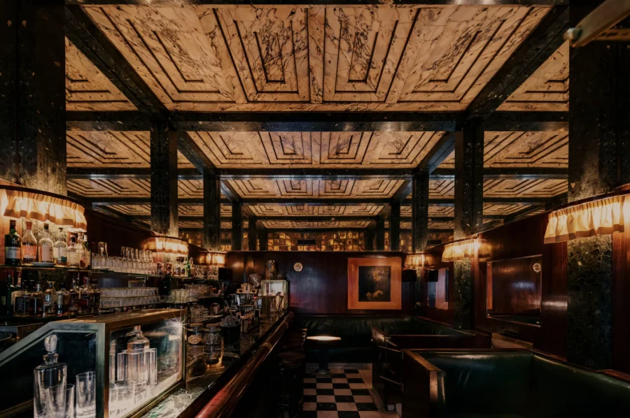 Dimly lit vintage bar interior with a marble patterned ceiling, wooden paneling, and green leather booths.