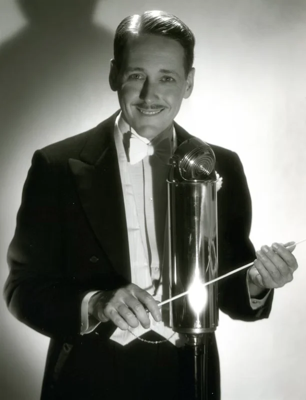 Black and white portrait of a man in a tuxedo holding a conductor's baton and standing behind a large vintage microphone.