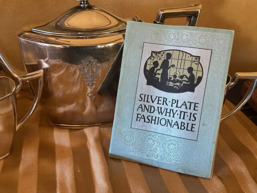 A silver tea set displayed next to a vintage book titled 'Silver Plate and Why It Is Fashionable' with an illustration of people drinking tea.