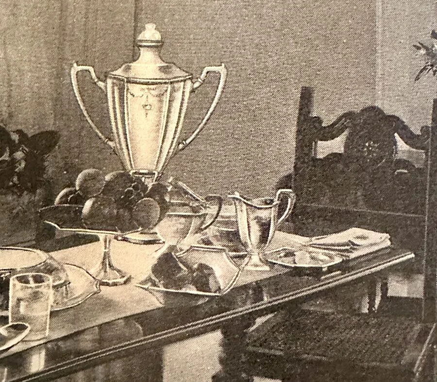 Vintage black and white photo of a dining table with silver tea set, fruit bowl, bread, glass of milk, and ornate wooden chairs.