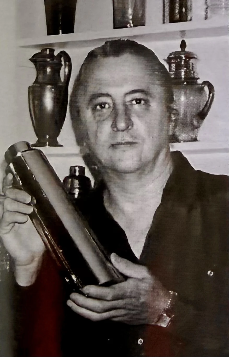 Black and white photo of a man holding a book or object, with trophies or decorative vessels on shelves behind him.