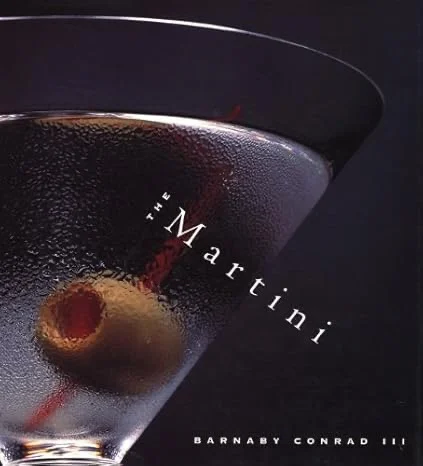 Close-up of a chilled martini glass with a green olive on a red cocktail pick, condensation visible on the glass surface.