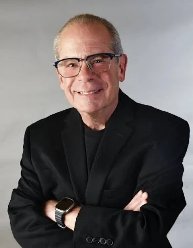 Smiling older man wearing glasses and a black blazer with arms crossed.