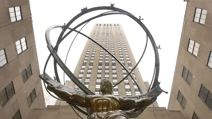 Low-angle view of a bronze statue holding large metal rings in front of a tall office building