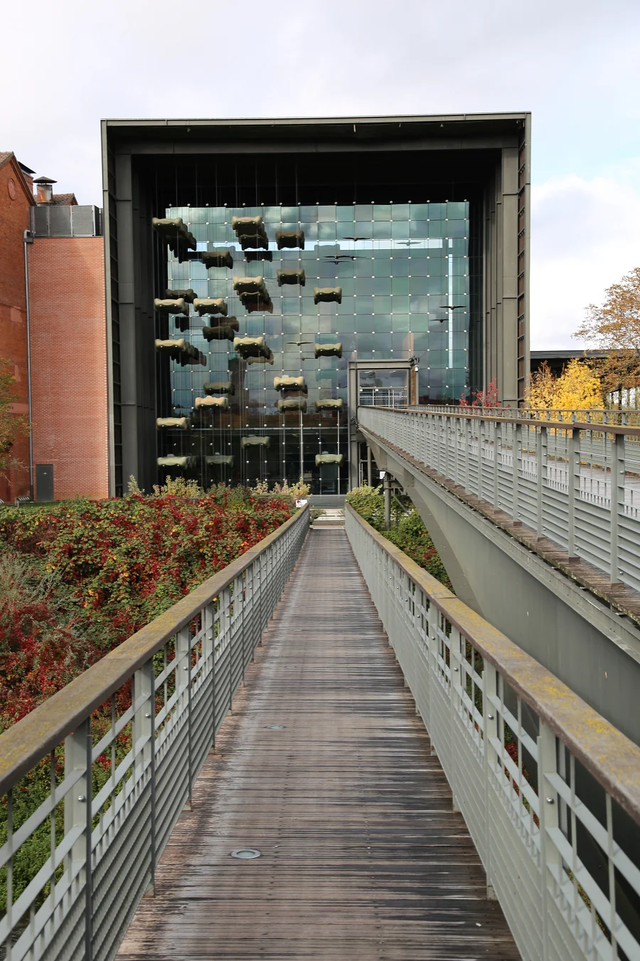 Modern glass building with hanging balconies viewed from a wooden pedestrian bridge